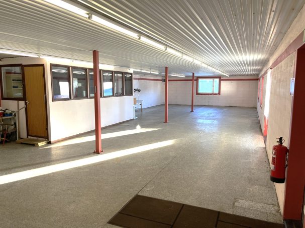 Tom lokal med vit vägg, betonggolv och fönster. Brandsläckare synlig.